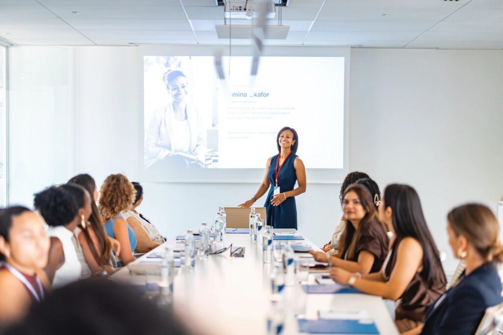 Female Speaker Standing At Conference Table While Presenting Slide To Seated Group Of Women During Business Conference Meeting In Bright Board Room